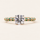 Round cut lab grown diamond engagement ring with green emerald gemstone accents, white diamond pavé shoulders, split shank and raised setting in yellow gold.