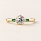 Round cut lab grown diamond bezel engagement ring with 0.50 ct center diamond, two tapered baguette green emerald side stones and polished yellow gold band.