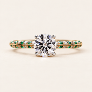 Round cut lab grown diamond engagement ring with green emerald gemstone accents, white diamond pavé shoulders, split shank and raised setting in yellow gold.