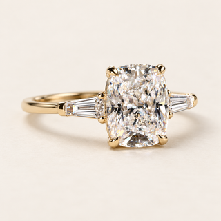 Cushion cut lab grown diamond three-stone engagement ring in yellow gold with four corner claws, tapered baguette side diamonds and a polished band.
