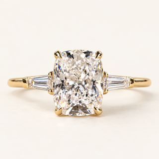 Cushion cut lab grown diamond three-stone engagement ring in yellow gold with four corner claws, tapered baguette side diamonds and a polished band.