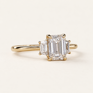 Emerald cut lab grown diamond three-stone engagement ring in yellow gold with trapezoid side diamonds, four center claws and a plain band