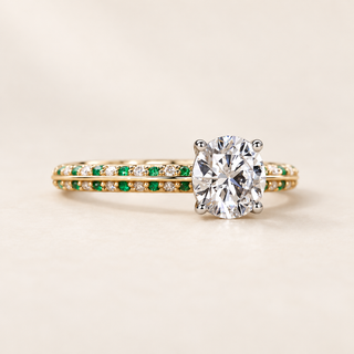 Round cut lab grown diamond engagement ring with green emerald gemstone accents, white diamond pavé shoulders, split shank and raised setting in yellow gold.
