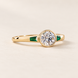 Round cut lab grown diamond bezel engagement ring with 0.50 ct center diamond, two tapered baguette green emerald side stones and polished yellow gold band.