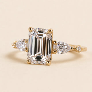 Emerald cut Lab Grown Diamond three stone engagement ring in yellow gold with pear side diamonds and four-prong basket setting.