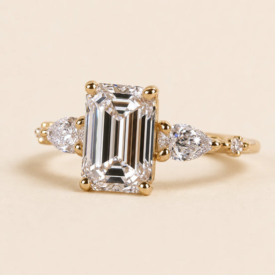Emerald cut Lab Grown Diamond three stone engagement ring in yellow gold with pear side diamonds and four-prong basket setting.