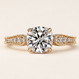 Round cut lab grown diamond engagement ring with six-claw center setting, round diamond-set leaf shoulders, milgrain edges and filigree detailing in yellow gold.