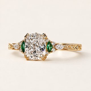 Cushion cut lab grown diamond engagement ring with green marquise emerald gemstones, round diamond accents, decorative filigree and milgrain yellow gold shoulders.