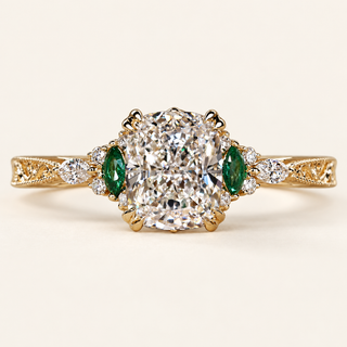 Cushion cut lab grown diamond engagement ring with green marquise emerald gemstones, round diamond accents, decorative filigree and milgrain yellow gold shoulders.