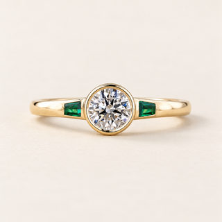 Round cut lab grown diamond bezel engagement ring with 0.50 ct center diamond, two tapered baguette green emerald side stones and polished yellow gold band.