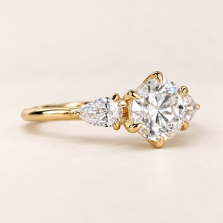 Round cut lab grown diamond three-stone engagement ring in yellow gold with six-prong center, pear-cut side diamonds and polished shank.