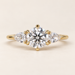 Round cut lab grown diamond three-stone engagement ring in yellow gold with six-prong center, pear-cut side diamonds and polished shank.